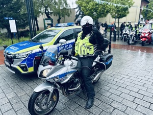 na zdjęciu umundurowany policjant ruchu drogowego siedzący na policyjnym motocyklu pokazuje ręką znak &quot;ok&quot;. Policjant stoi obok policyjnego radiowozu, a za nim motocykliści na motorach.