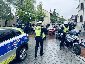 na zdjęciu oznakowany radiowóz policyjny, a obok niego umundurowany policjant, który idzie w stronę motocyklistów.